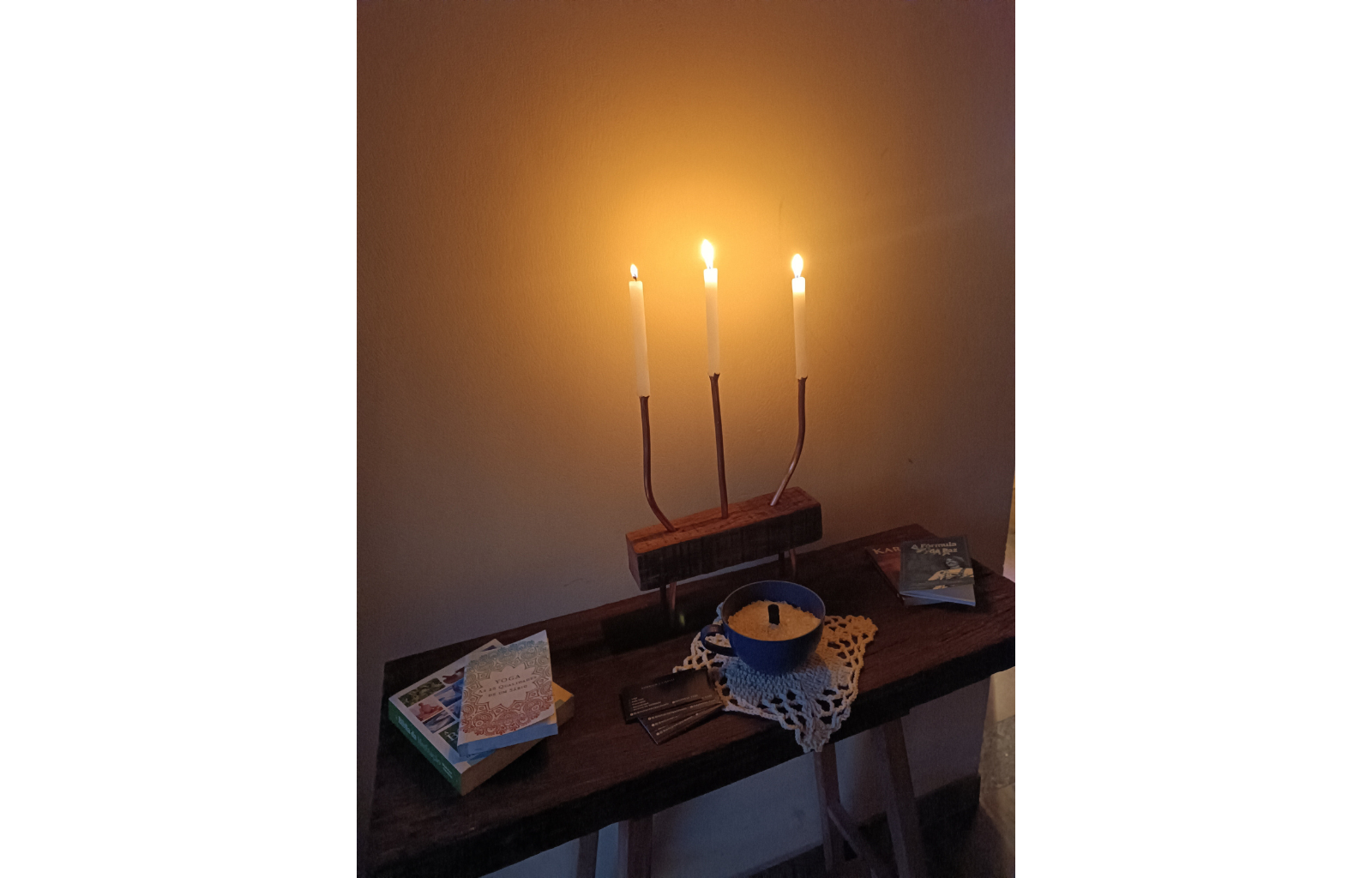 Imagem de um castiçal com três velas acesas em um aparador. Neste aparador encontra-se livros de bolso e cartões de visita do terapeuta Rodrigo Cainelli.