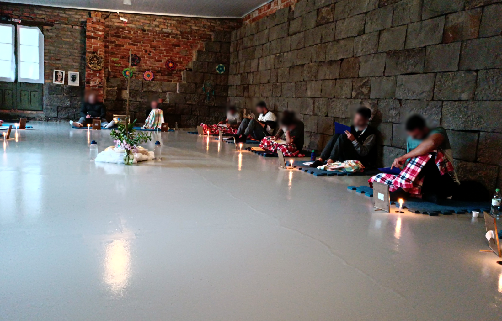 Imagem registrada de uma imersão para homens promovida pelo terapeuta Rodrigo. Nesta imagem os homens estão sentados sobre um tatame, alguns estão utilizando uma pequena manta para se aquecer e todos estão tomando nota em um caderno de anotações. Este registro mostra a outra metade dos participantes.