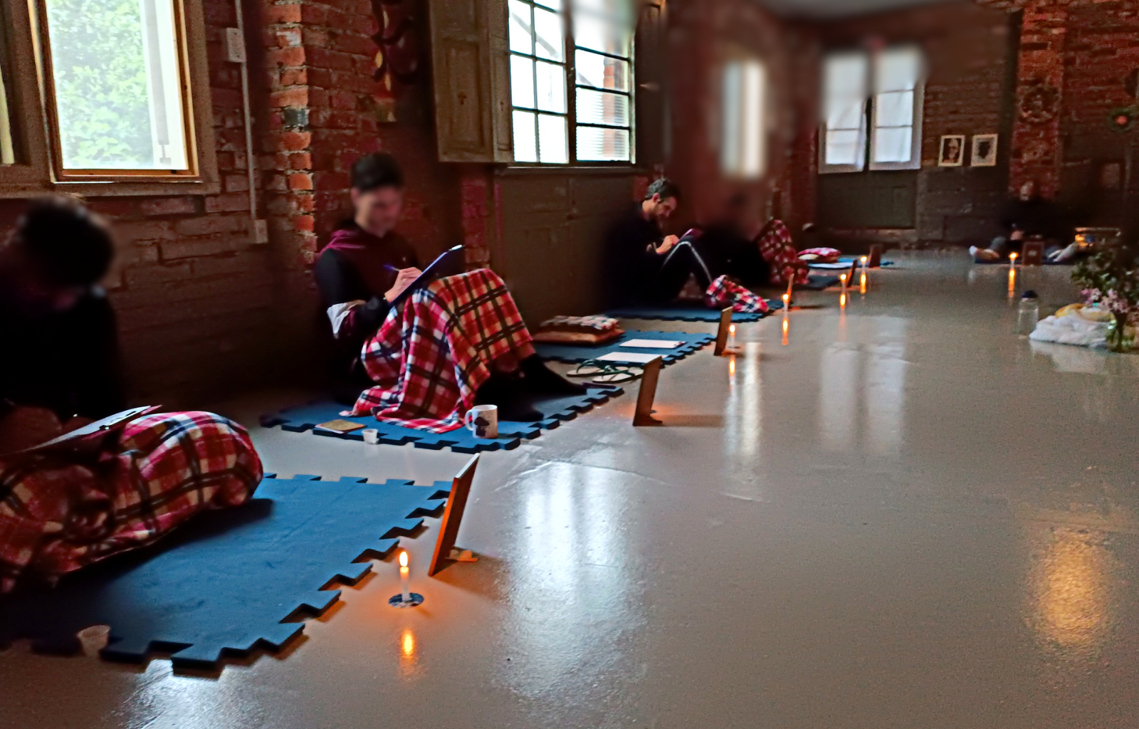 Imagem registrada de uma imersão para homens promovida pelo terapeuta Rodrigo. Nesta imagem os homens estão sentados sobre um tatame, alguns estão utilizando uma pequena manta para se aquecer e todos estão tomando nota em um caderno de anotações. Este registro mostra metade dos participantes.